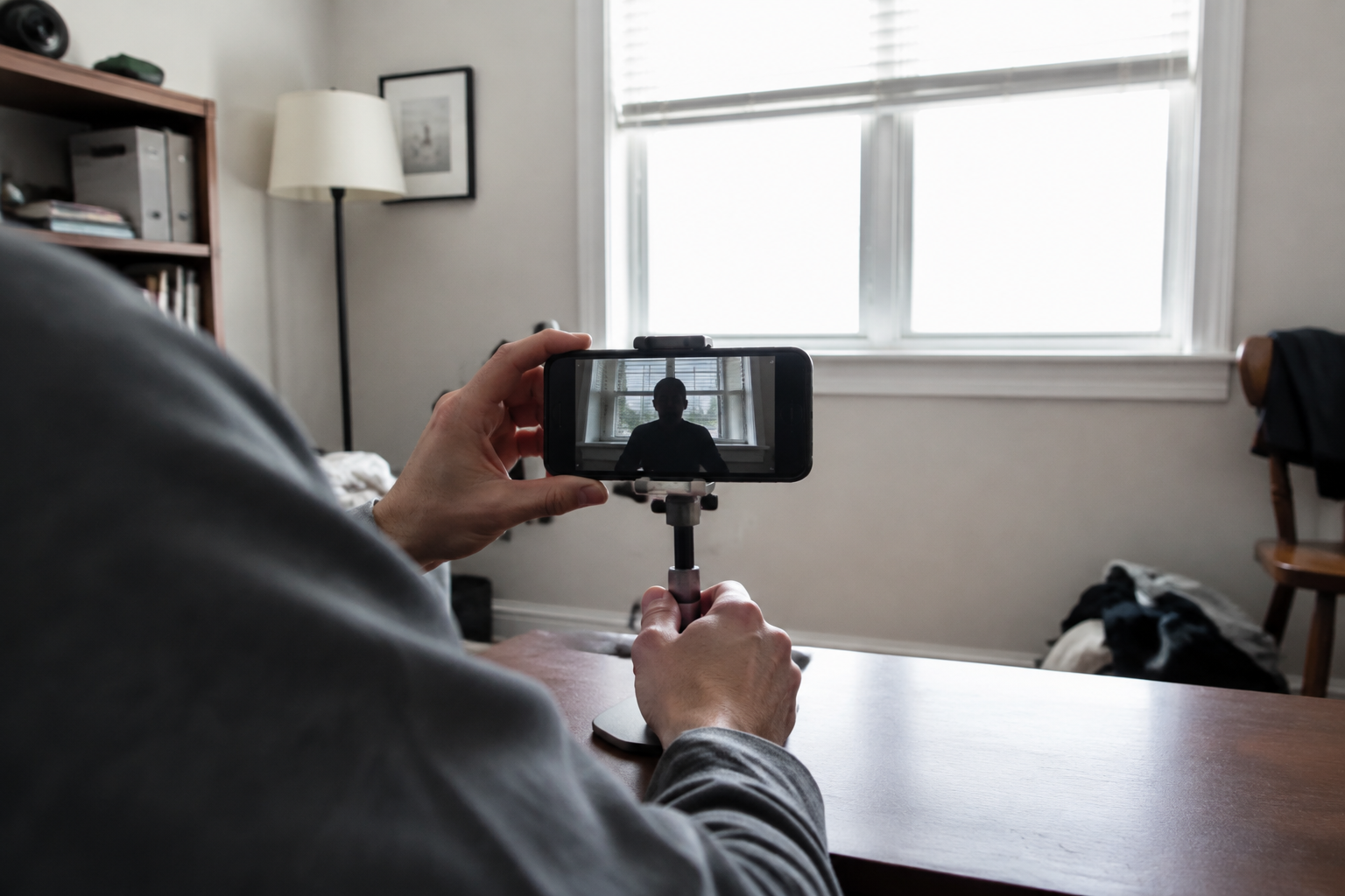 Over-the-shoulder view of a phone recording a dark silhouette against a bright window in a modest home office.