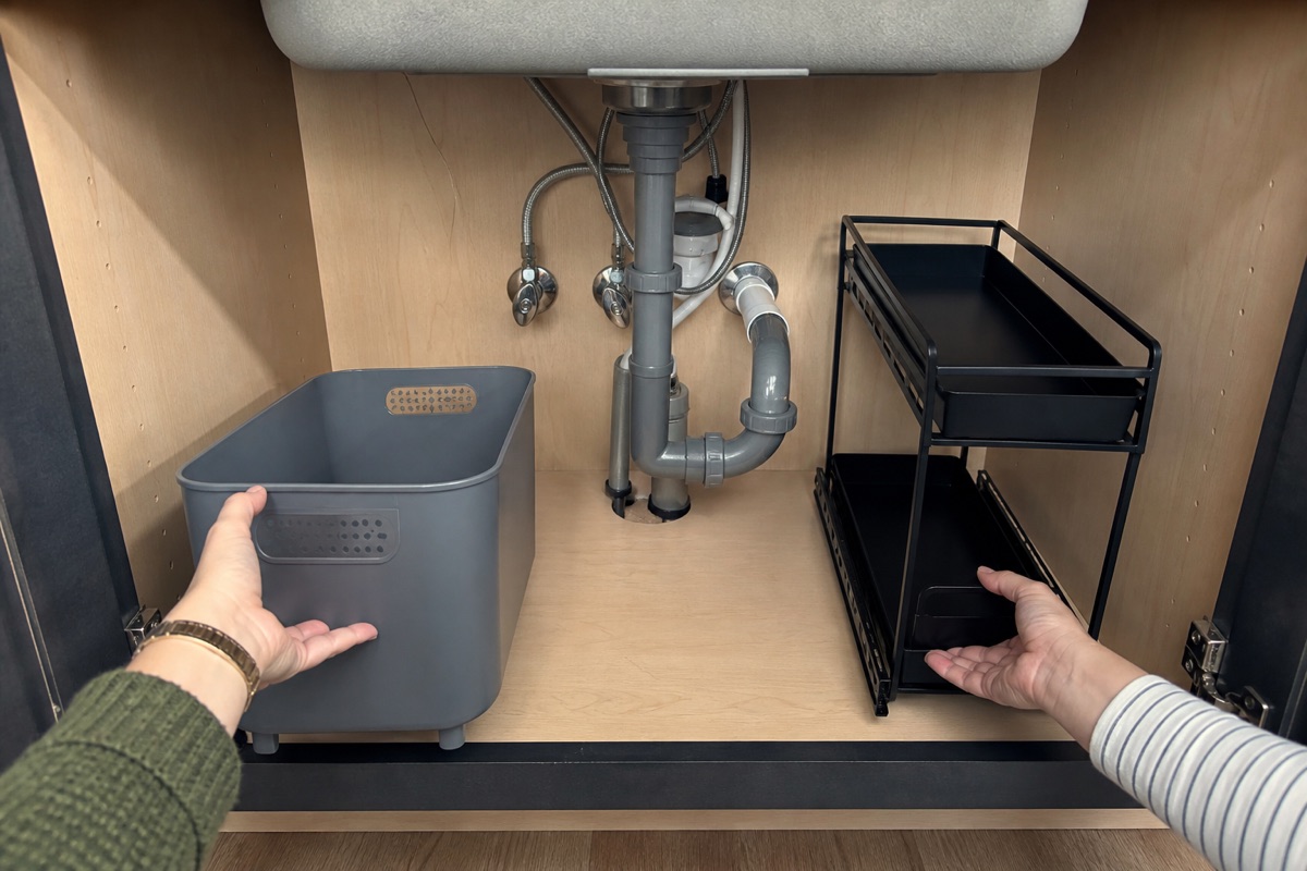 Hands compare a removable bin with a two-tier organizer on opposite sides of visible under-sink plumbing.