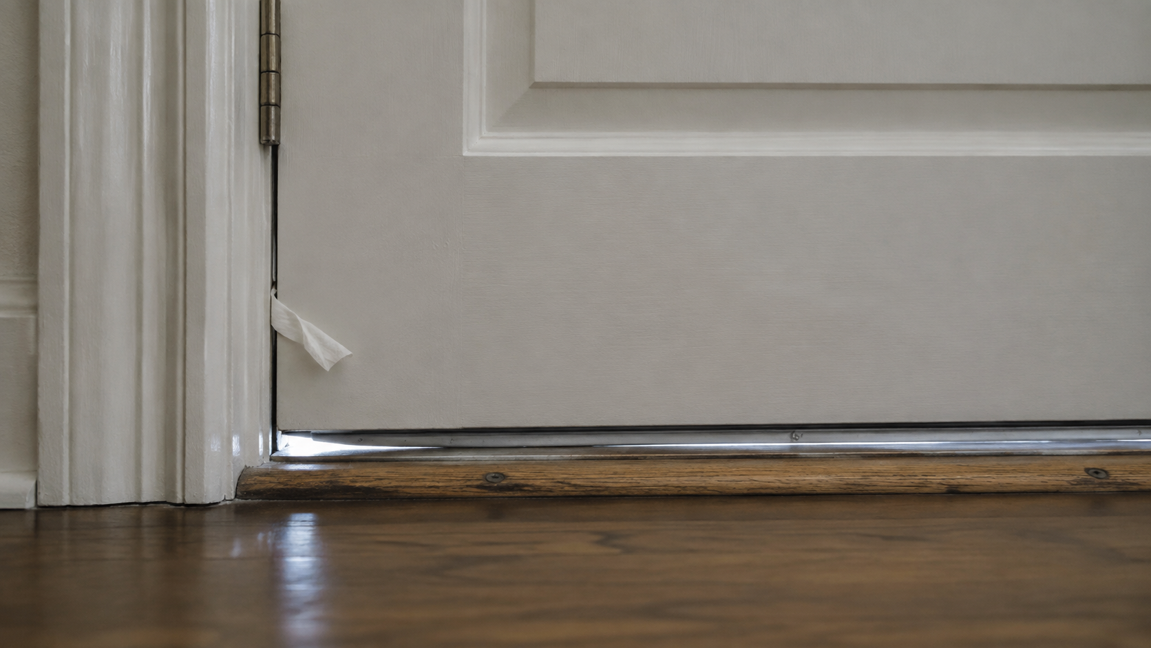A closed exterior door in an ordinary home with a small visible corner gap and a lightweight tissue used to locate air movement along the jamb.