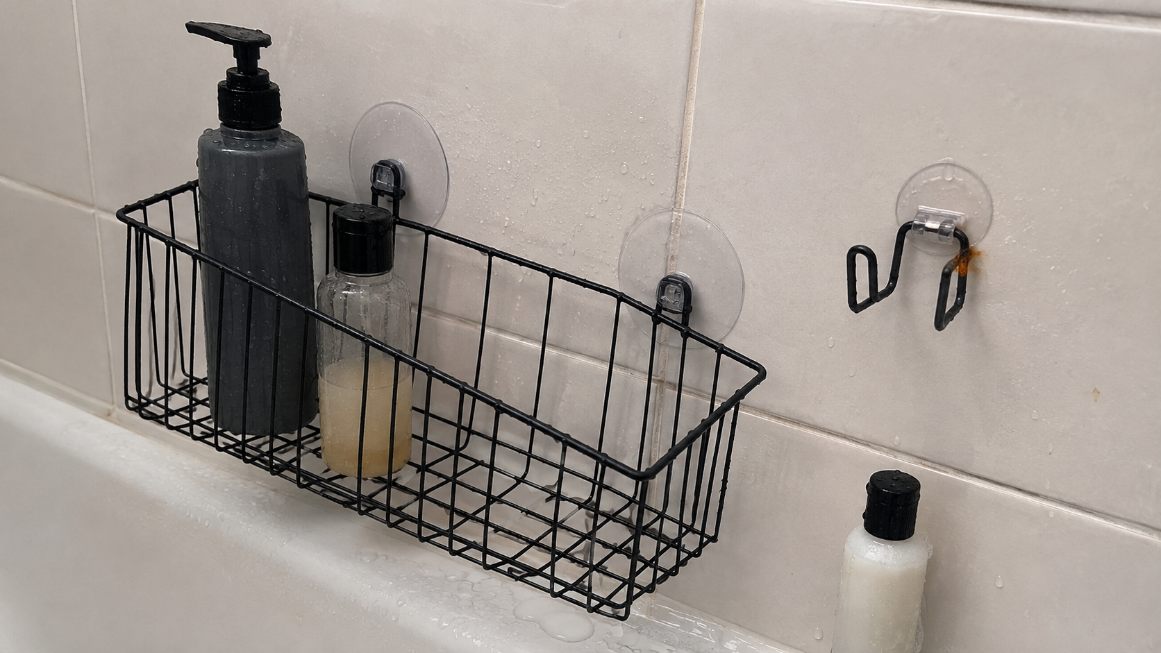A black shower basket with two suction cups, a loose mounting hook, and a small rust-colored spot beside a bottle in an ordinary tiled shower.
