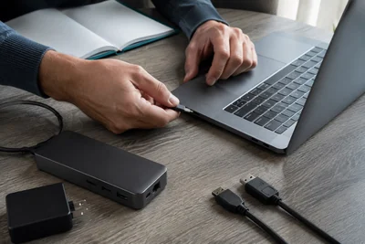 AI illustration of hands checking a USB-C connection beside a laptop, dock and charging equipment.