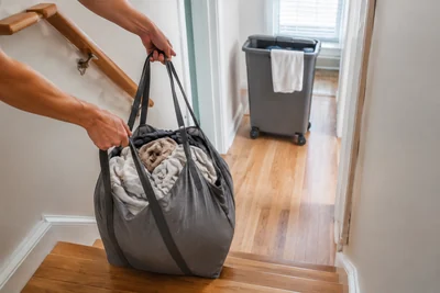 Hands carrying a soft-sided laundry hamper at the top of a staircase while a rigid wheeled hamper remains on the flat hallway floor.