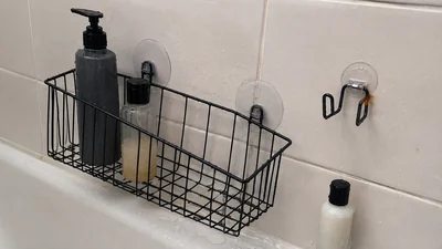 A black shower basket with two suction cups, a loose mounting hook, and a small rust-colored spot beside a bottle in an ordinary tiled shower.