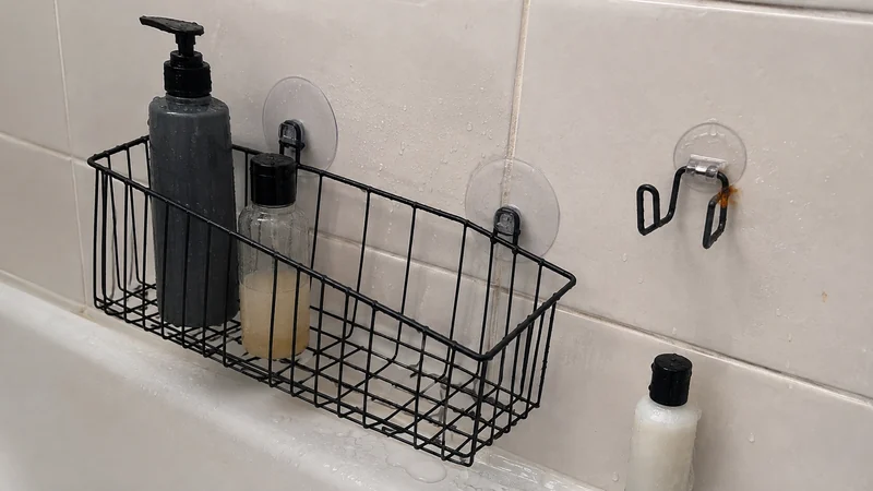 A black shower basket with two suction cups, a loose mounting hook, and a small rust-colored spot beside a bottle in an ordinary tiled shower.