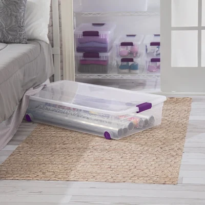 Clear Sterilite 60-quart wheeled under-bed storage box with white lid and purple latches beside a bed.