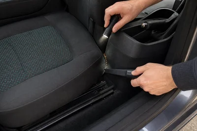 Hands using a flashlight and crevice nozzle to inspect dry debris between a car seat and console.